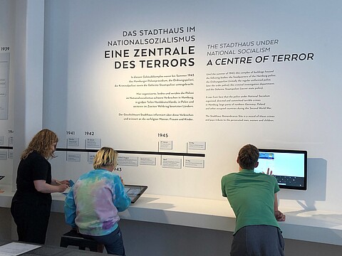 Drei Personen stehen vor einer Ausstellung und sehen sich etwas auf zwei Bildschirmen an. 