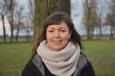 Malina trägt einen großen Wollschal und eine Jacke. Im Hintergrund Wiese und Bäume